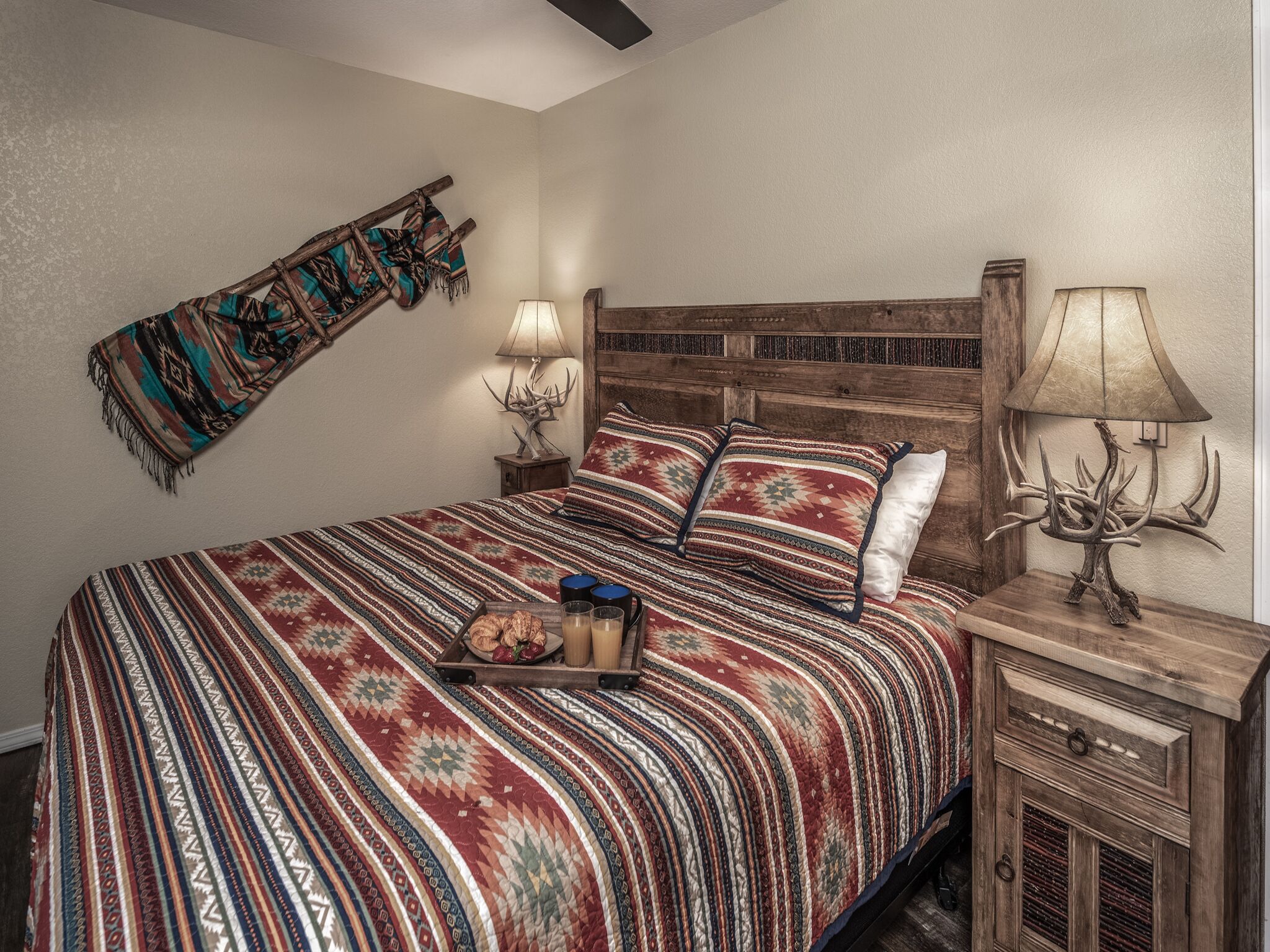 A bedroom with a large bed, a wooden headboard, two bedside tables with lamps, a blanket on the wall, and a small wooden cabinet.