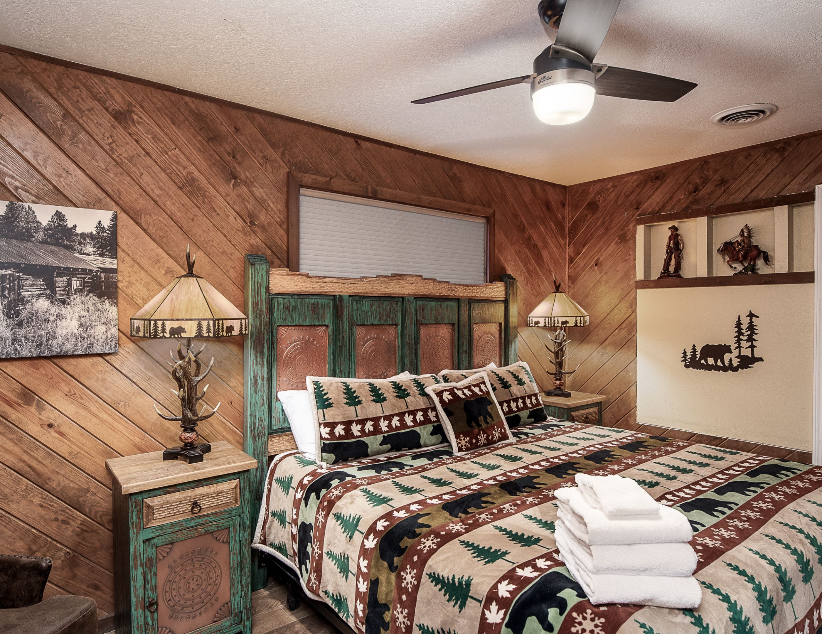 Une chambre à coucher au style rustique, avec un mur lambrissé en bois, un lit recouvert d’une couvre-lit à motifs, deux lampes de chevet et un ventilateur de plafond.