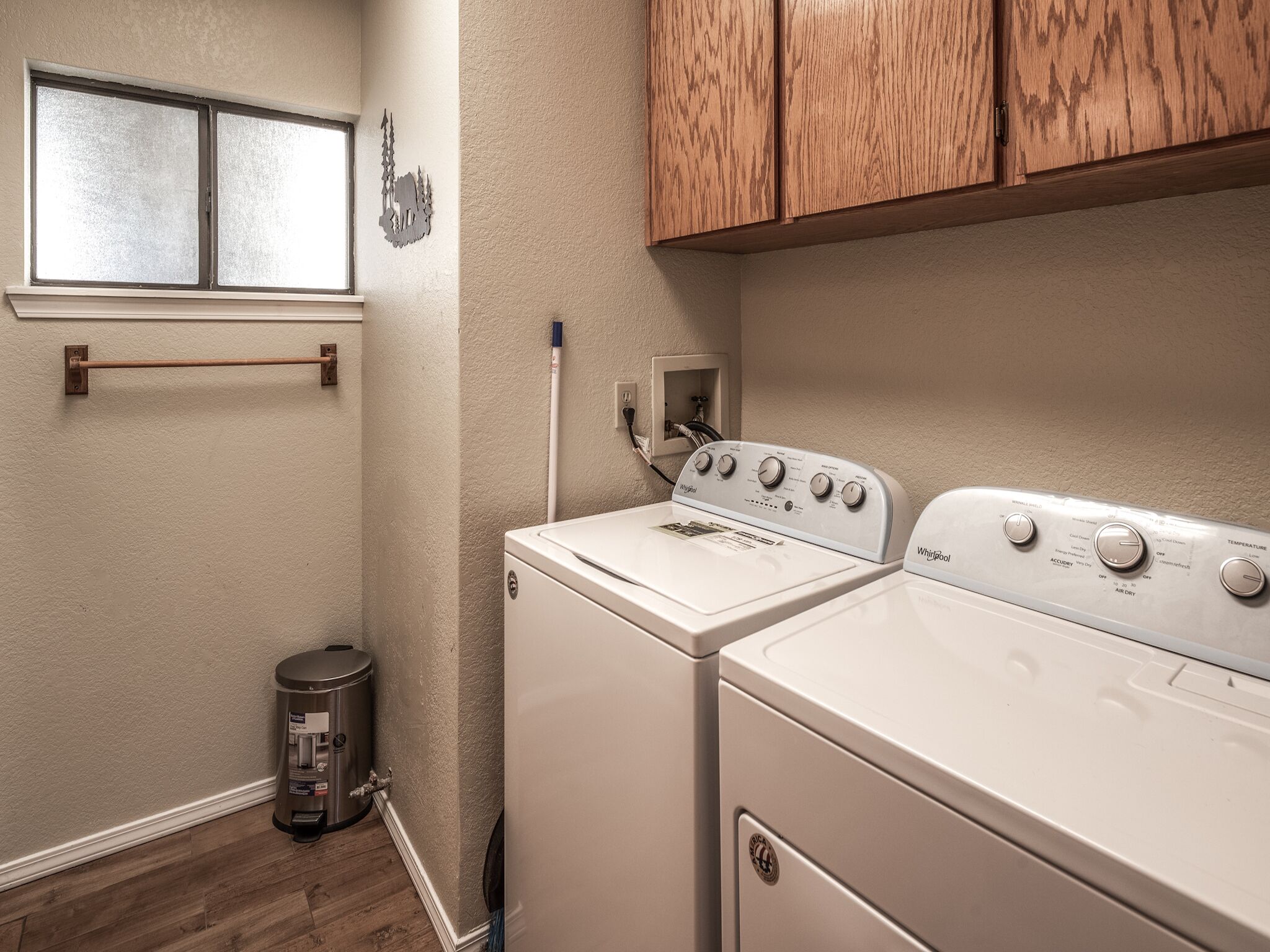 A laundry room with a front-loading washer and dryer, a small trash can, and wooden cabinets above the appliances.