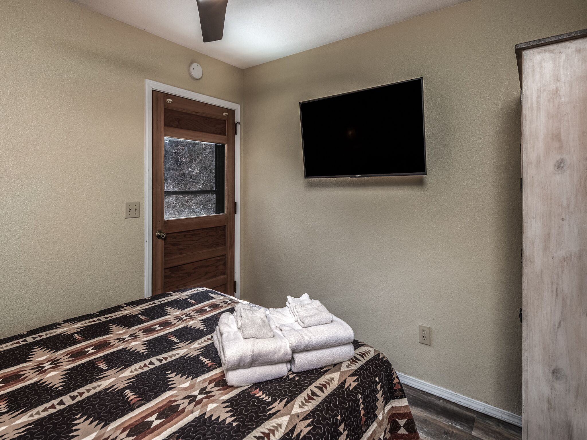 Un dormitorio con cama, televisor montado en la pared, puerta con ventana y armario de madera.