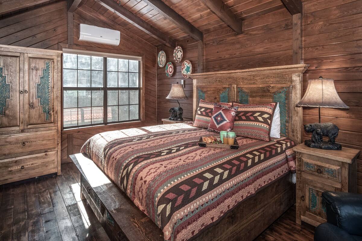 A rustic wooden bedroom with a large bed, a window, a dresser, and a nightstand with a lamp and a bear statue.