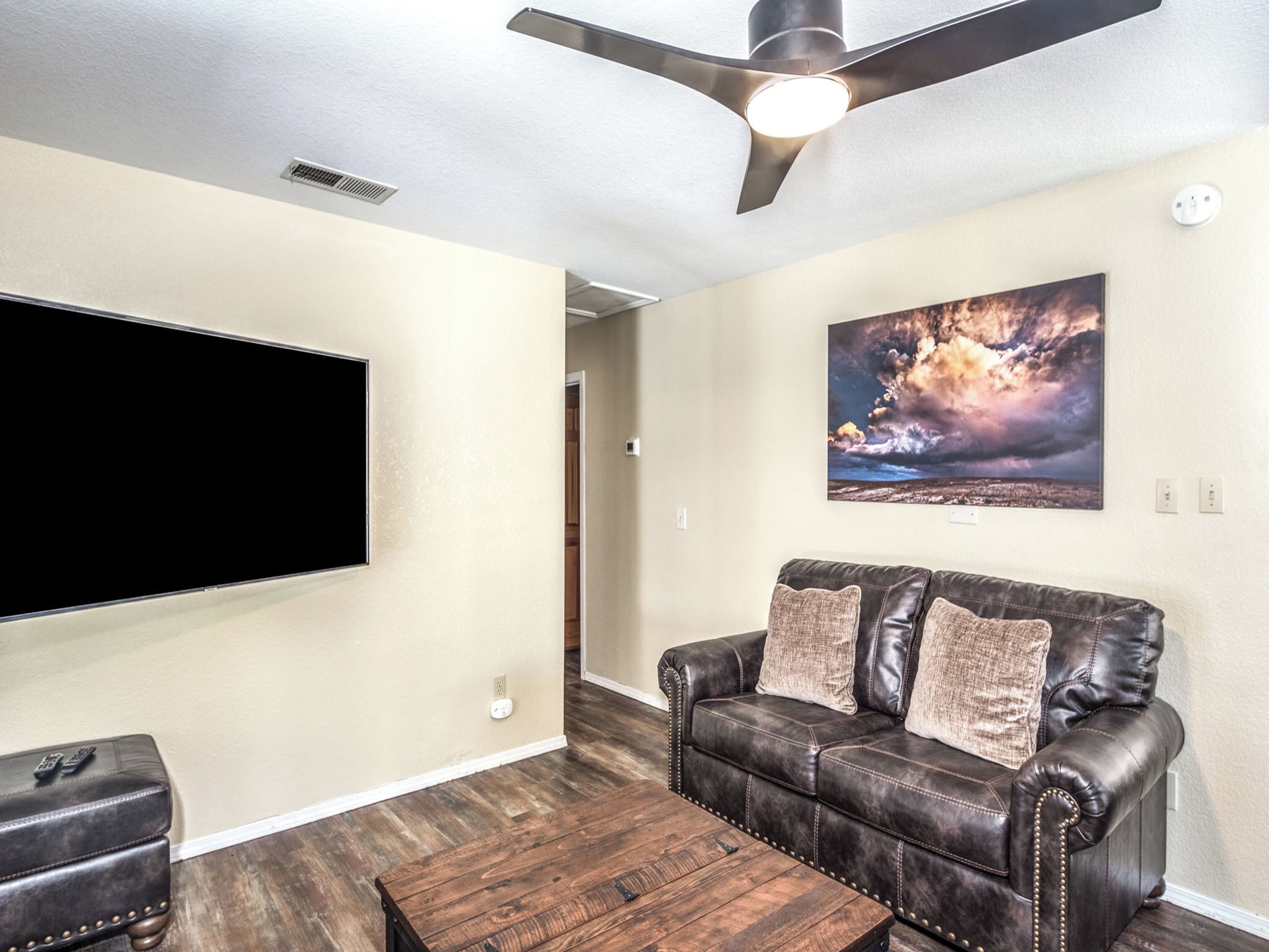 A living room with a leather sofa, a coffee table, a flat-screen TV mounted on the wall, and a ceiling fan.