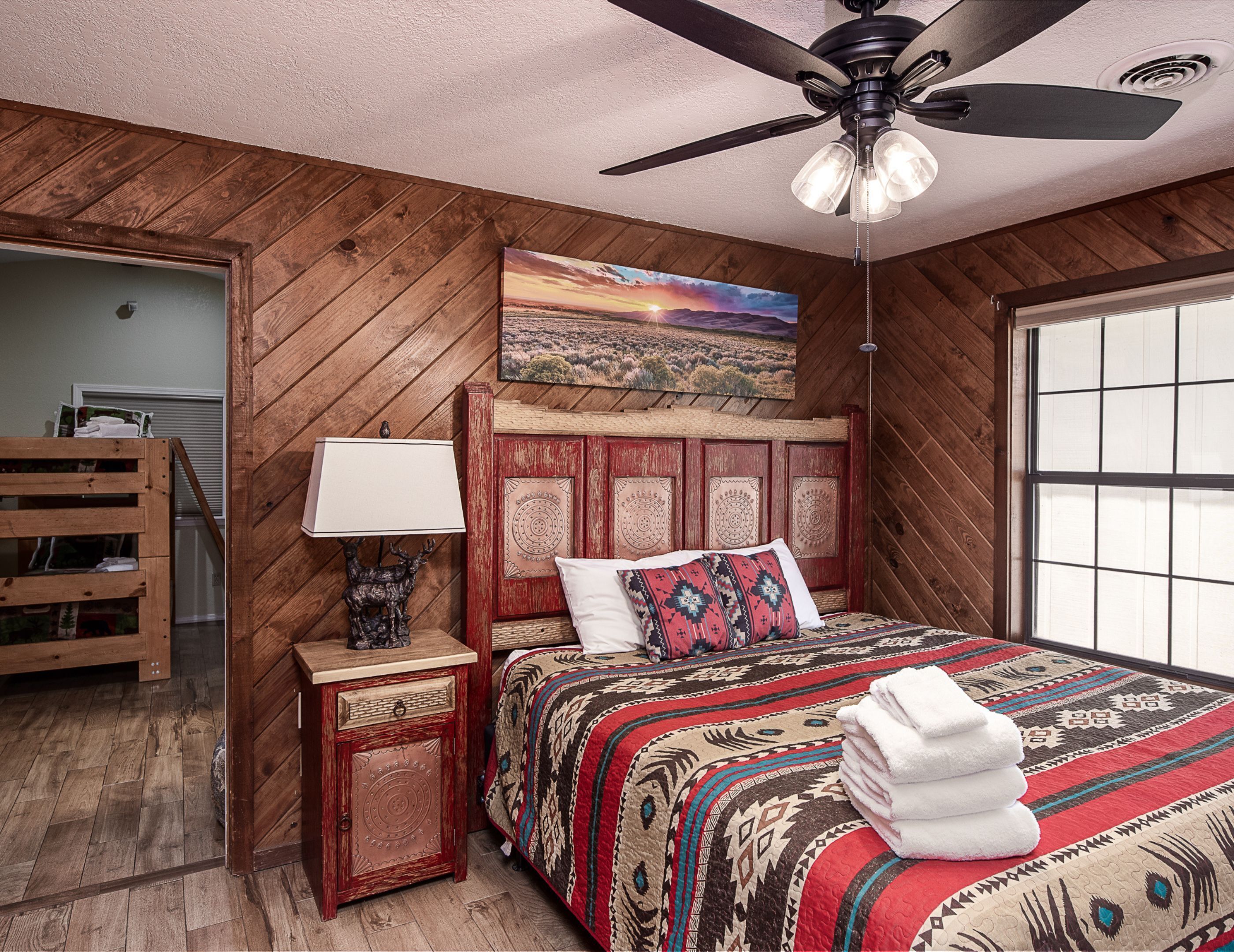 A bedroom with a wooden ceiling, a bed with patterned bedding, a nightstand with a lamp, and a painting above the bed.