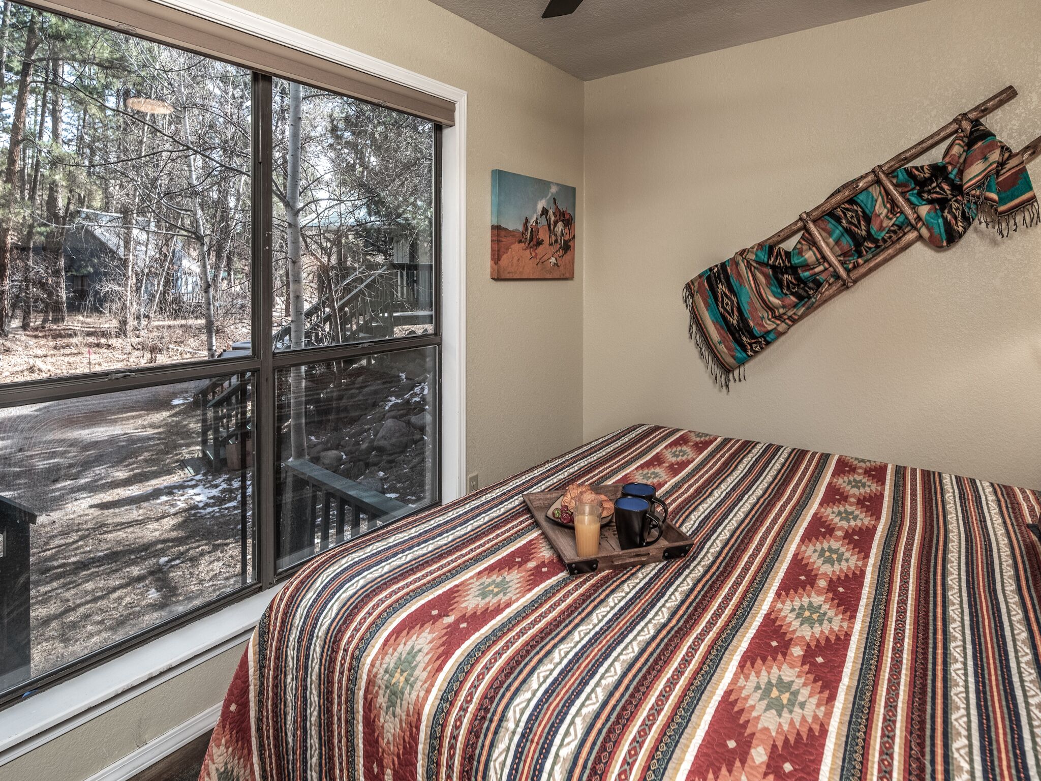 Ein Schlafzimmer mit großem Fenster, einem Bett mit gemustertem Überwurf, einem Bild an der Wand und einer Decke, die an der Wand hängt.