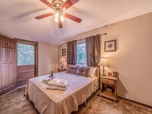 Quarto de hotel com uma cama grande, um ventilador de teto, uma janela com cortinas, uma mesa de cabeceira com abajur e um quadro na parede.
