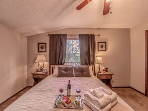 Quarto com cama grande, duas mesas de cabeceira com luminárias, janela com cortinas, ventoinha no teto e bandeja com comida e bebidas sobre a cama.