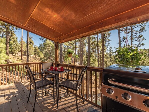 A covered outdoor area with a grill, a table with chairs, and a view of a forest. - My Happy Place by Ruidoso Vacation Rentals (Ruidoso)