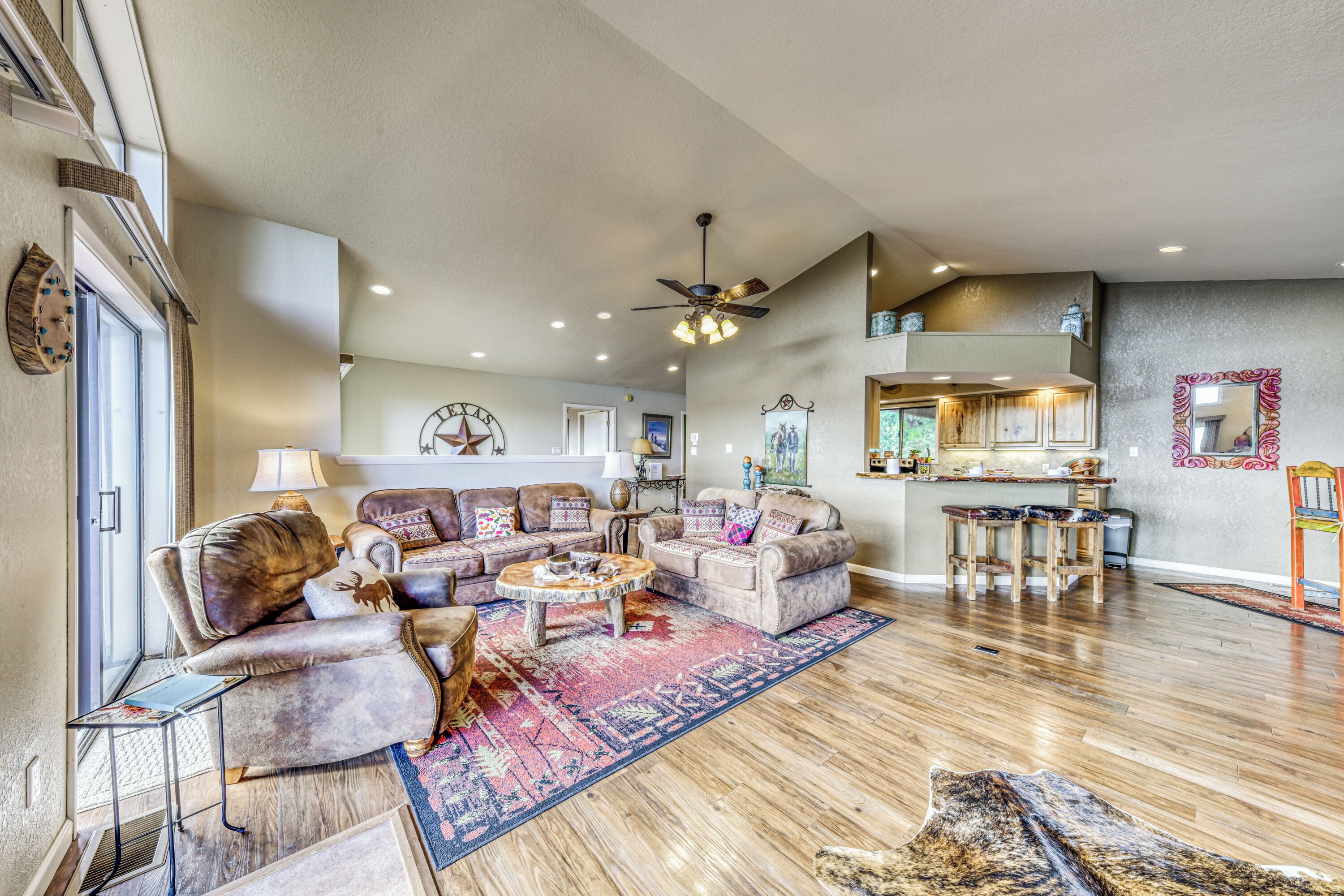 A spacious living room with a large sofa set, a coffee table, and a rug. There is a dining area with a table and chairs, and a kitchen in the background.