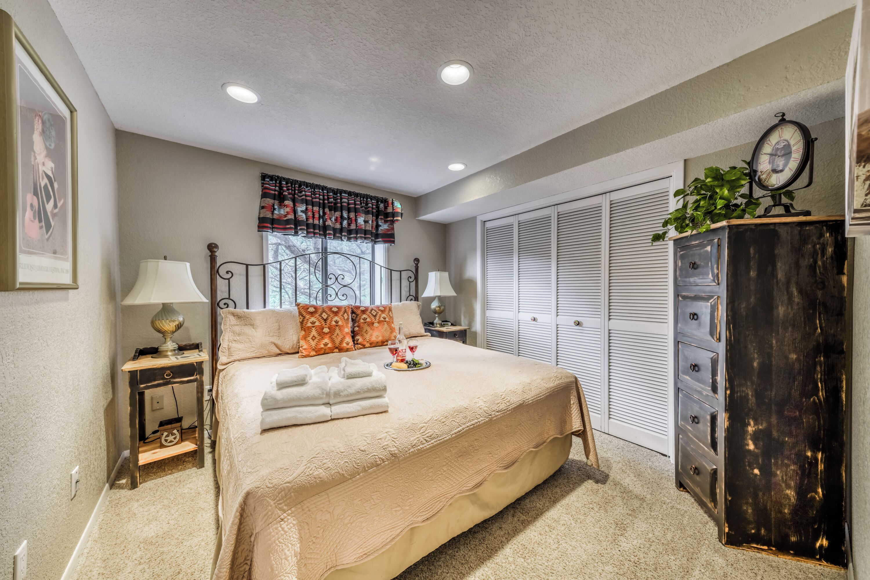 A bedroom with a large bed, two bedside tables with lamps, a dresser, a window with curtains, and a closet with louvered doors.
