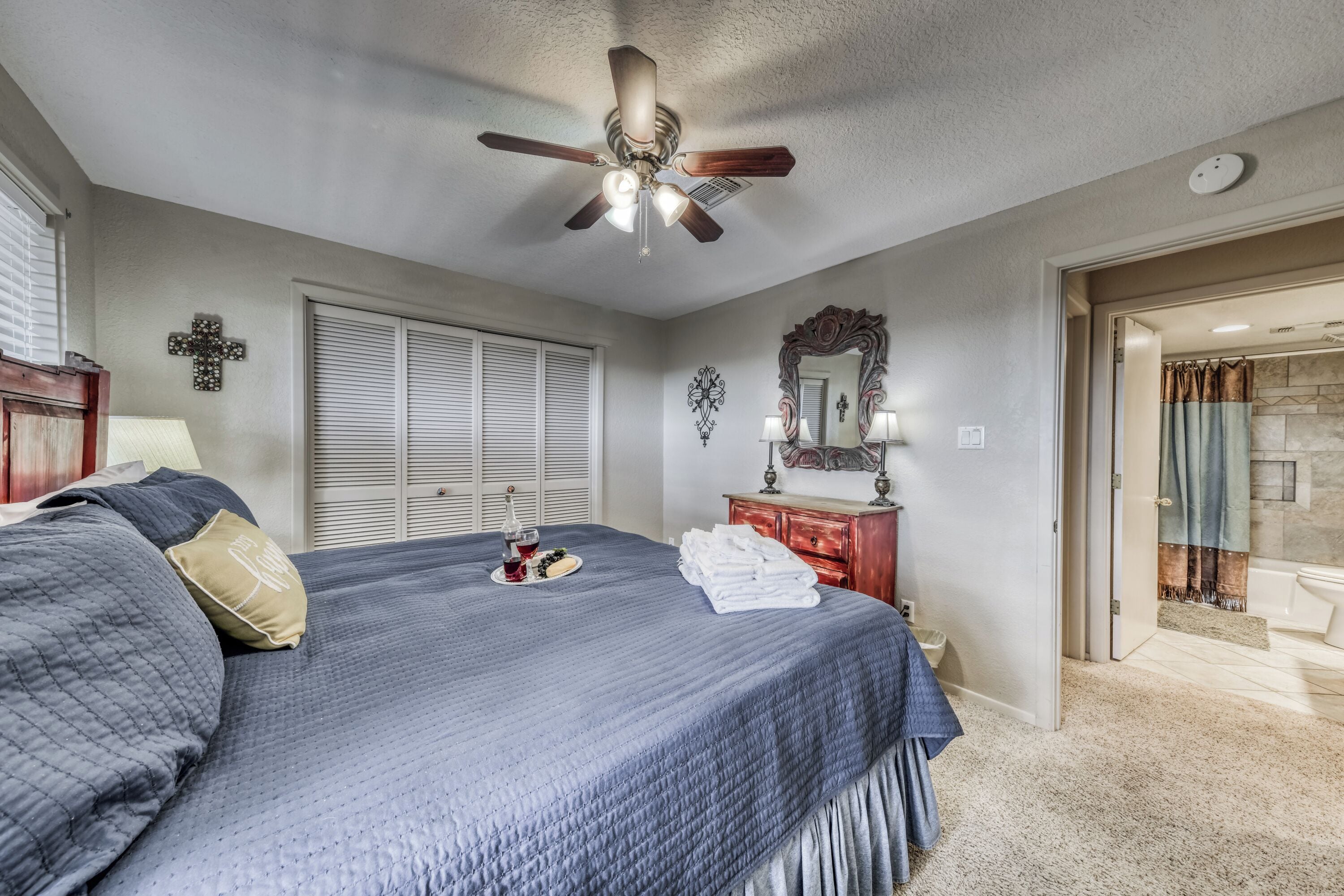 Une chambre avec un grand lit, un ventilateur de plafond, une commode, un miroir et une salle de bain visible à travers une porte ouverte.