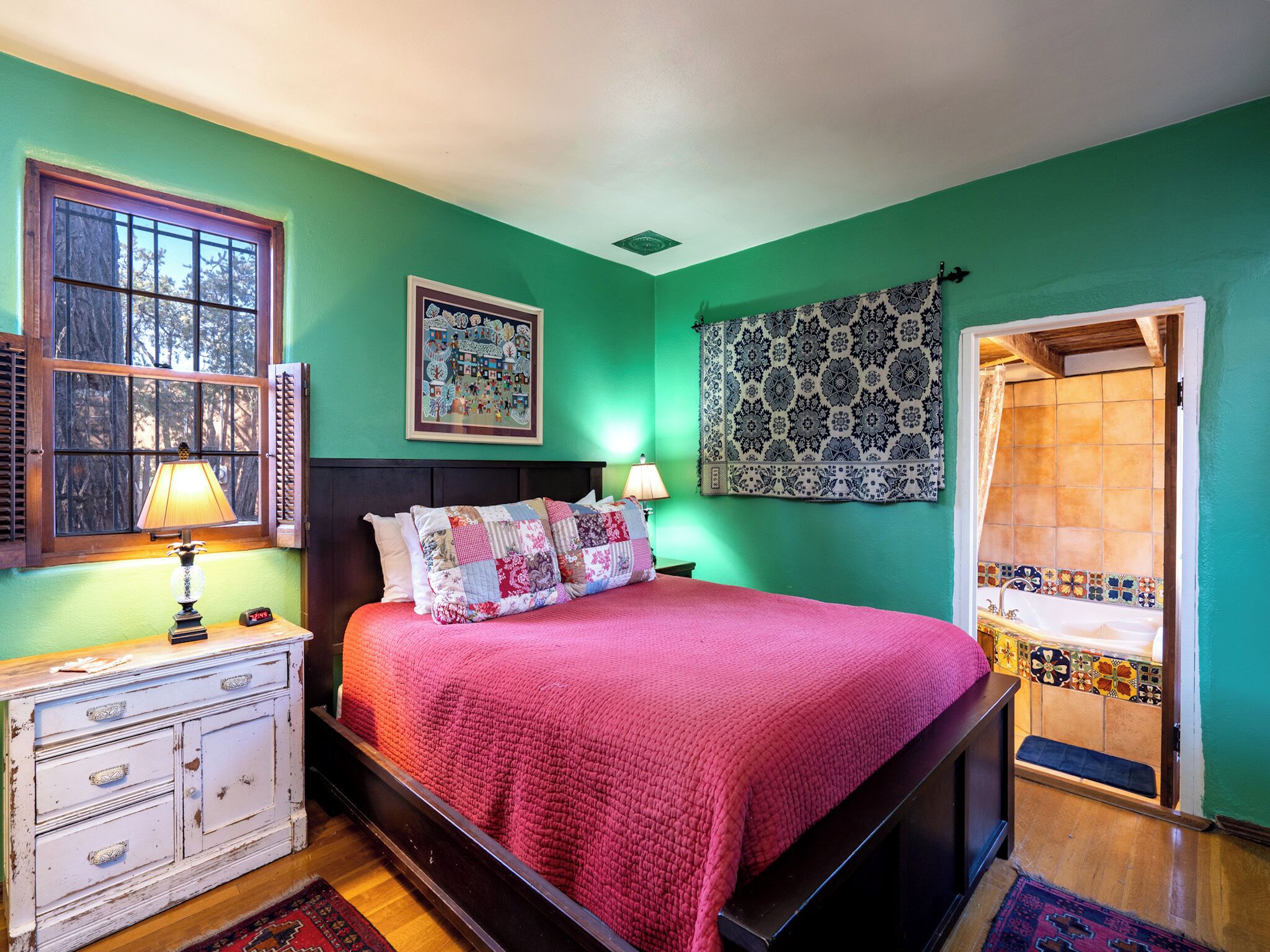 A bedroom with a bed, two bedside lamps, a dresser, a window with shutters, a patterned tapestry, and a bathroom visible through an open door.