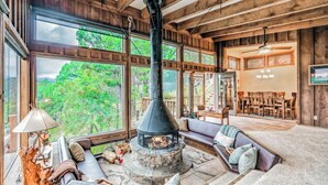 A cozy living room with a stone fireplace, large windows, and a wooden ceiling. - Neeley Mountain House by Ruidoso Vacation Rentals (Alto)