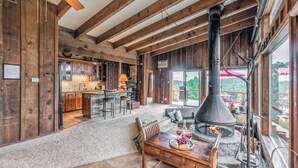 A rustic interior with exposed wooden beams, a stone fireplace, and a large window overlooking a deck. - Neeley Mountain House by Ruidoso Vacation Rentals (Alto)