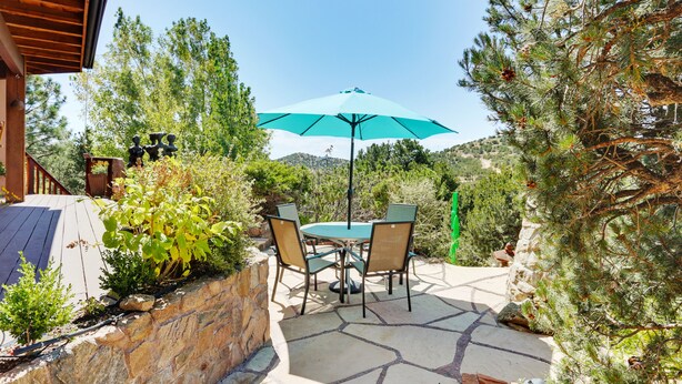 A patio with a table and chairs, a turquoise umbrella, and a stone wall. - Sunlit Hills Art and Views by Kokopelli Property Management (Santa Fe)