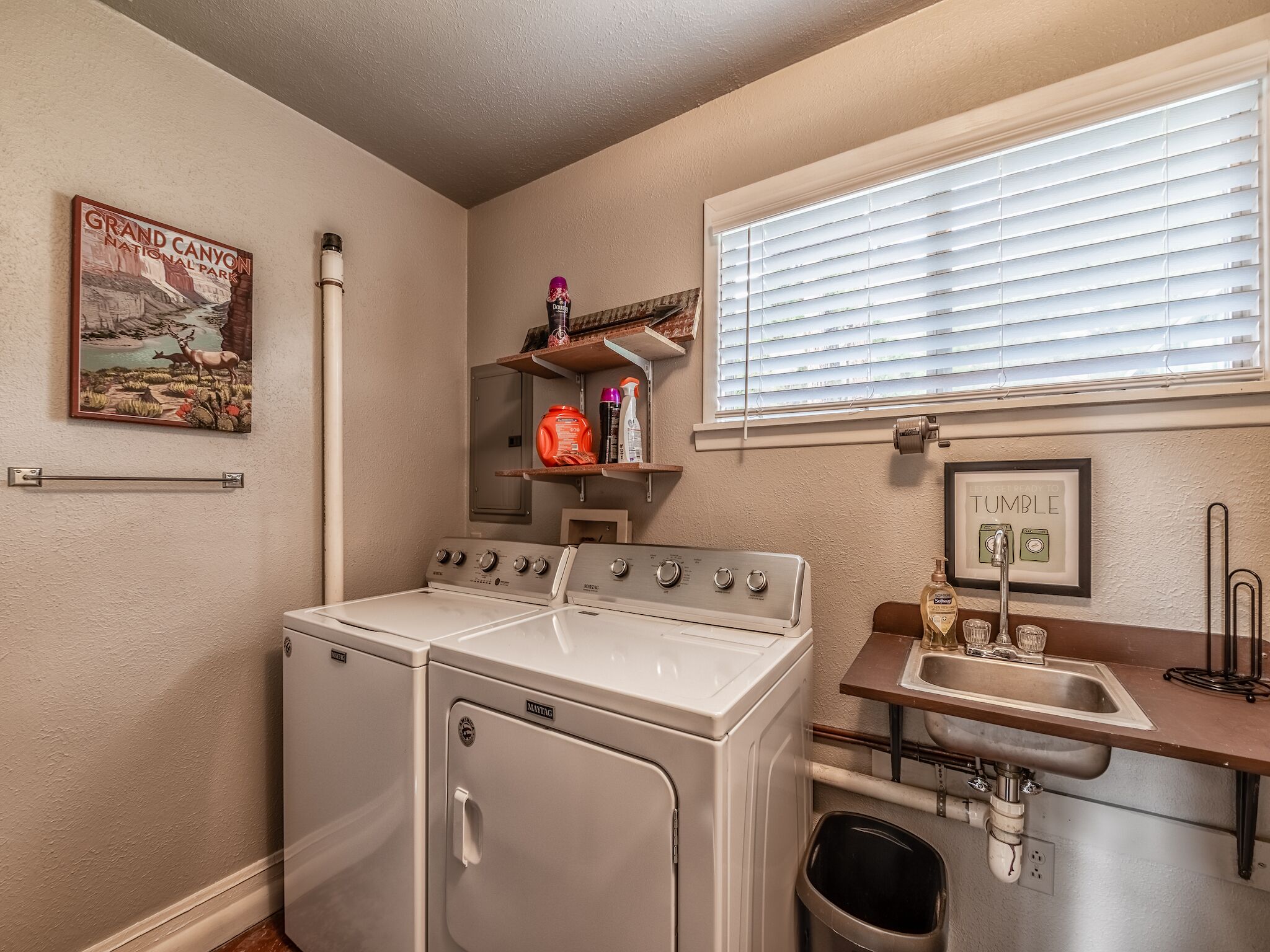 Un cuarto de lavado con lavadora y secadora Maytag, una ventana con persianas, un estante con artículos de aseo personal y un cartel del Gran Cañón.