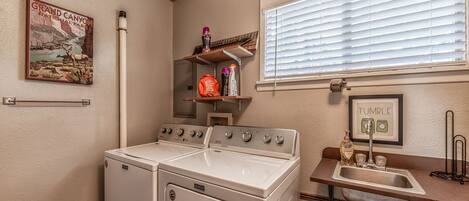 Un cuarto de lavado con lavadora y secadora Maytag, una ventana con persianas, un estante con artículos de aseo personal y un cartel del Gran Cañón.