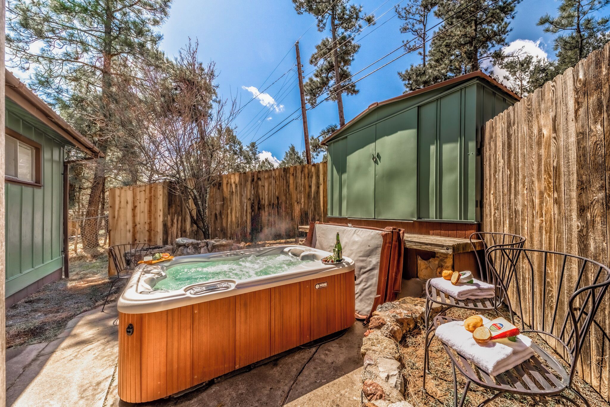 A hot tub in a backyard with a wooden fence, a green shed, and a metal chair.
