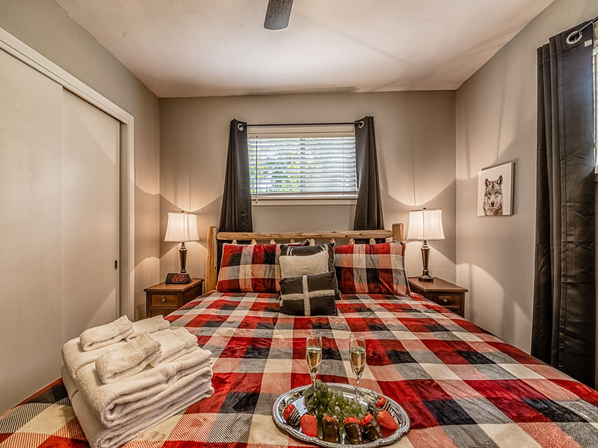 A hotel room with a checkered bedspread, two bedside lamps, a digital clock, a window with curtains, a framed picture on the wall, and a tray with grapes and strawberries.