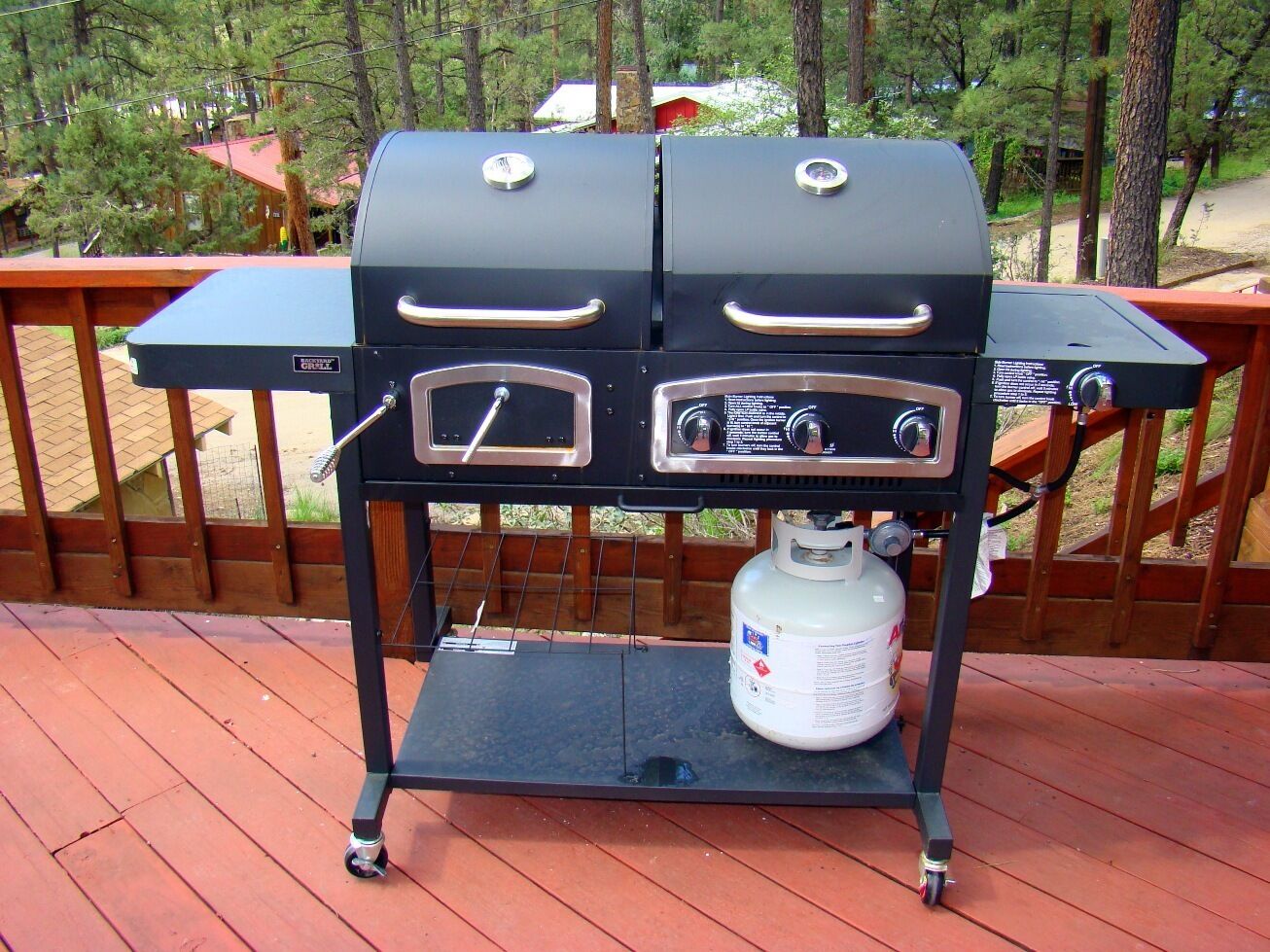 Ein schwarzer Grill mit zwei Kochzonen und einer Propangasflasche auf einer Holzterrasse.
