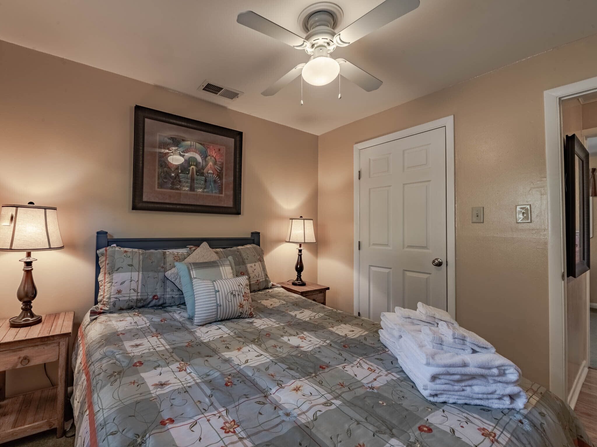 A bedroom with a bed, two bedside lamps, a ceiling fan, a door, and a framed picture on the wall.