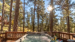 Ein Whirlpool auf einer Holzterrasse, umgeben von hohen Kiefern, mit blauem Himmel und durchschimmerndem Sonnenlicht.