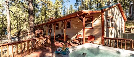 Una cabaña con jacuzzi, terraza de madera y zona de estar al aire libre, rodeada de árboles.