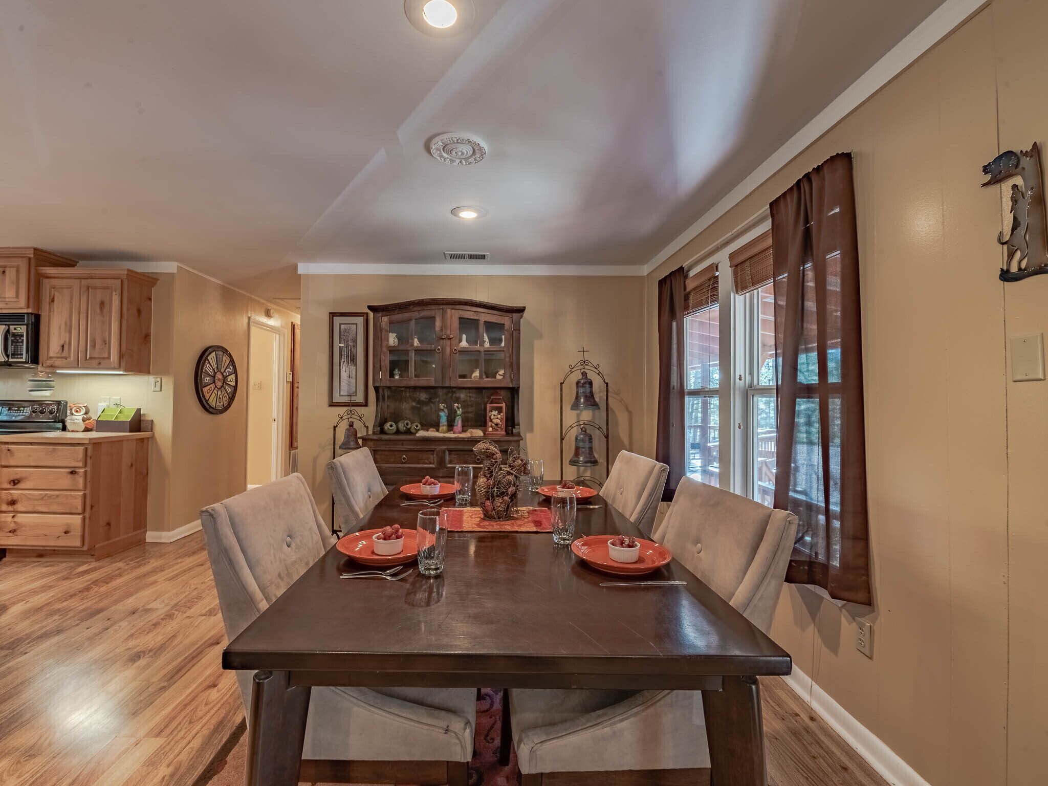 A dining area with a wooden table set for four, surrounded by beige chairs. A wooden cabinet with glass doors is in the background, and a decorative bell is mounted on the wall.