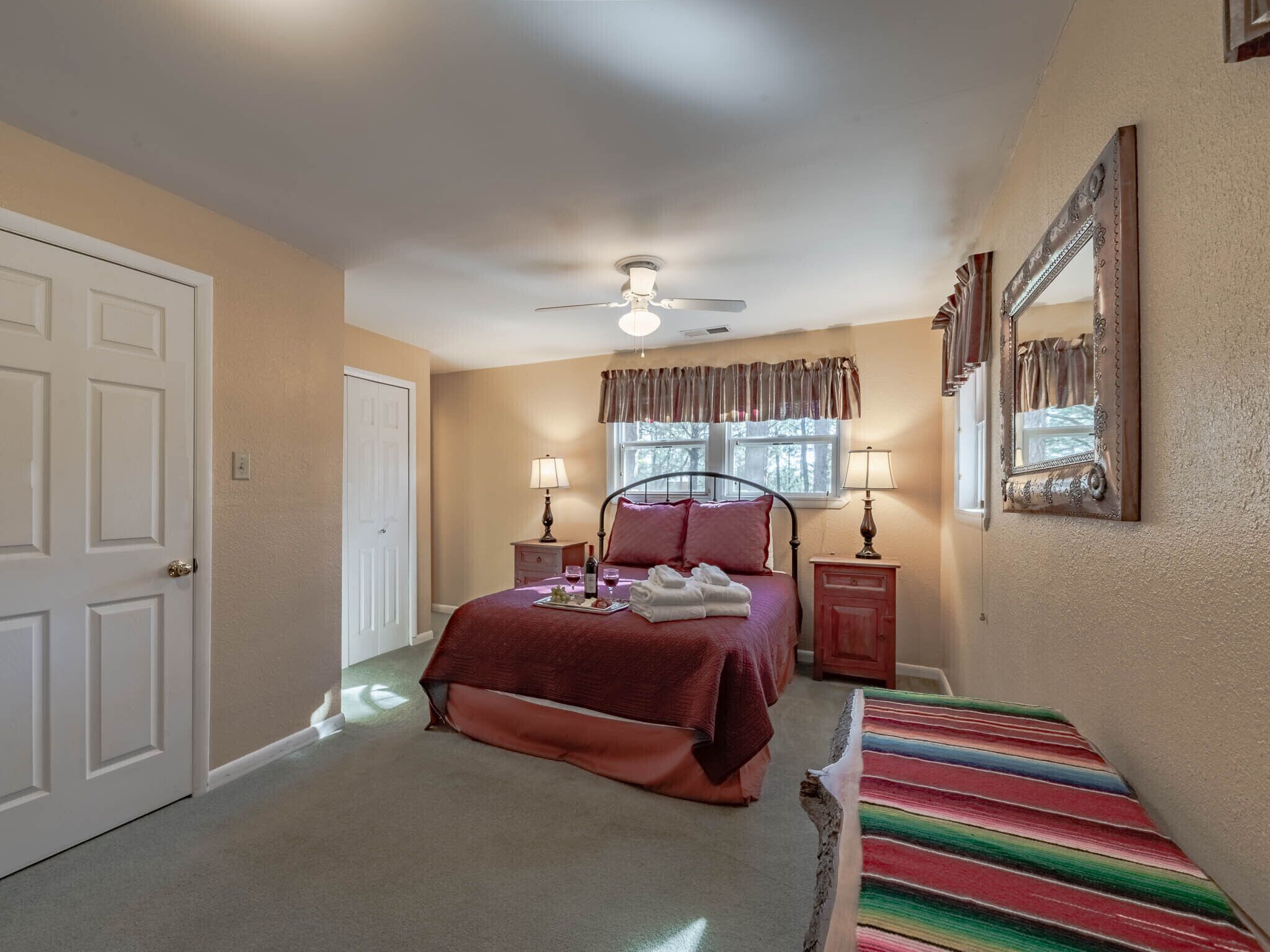 A bedroom with a bed, two bedside tables, a ceiling fan, a window with curtains, a mirror, and a colorful striped rug.