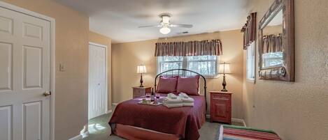 A bedroom with a bed, two bedside tables, a ceiling fan, a window with curtains, a mirror, and a colorful striped rug.