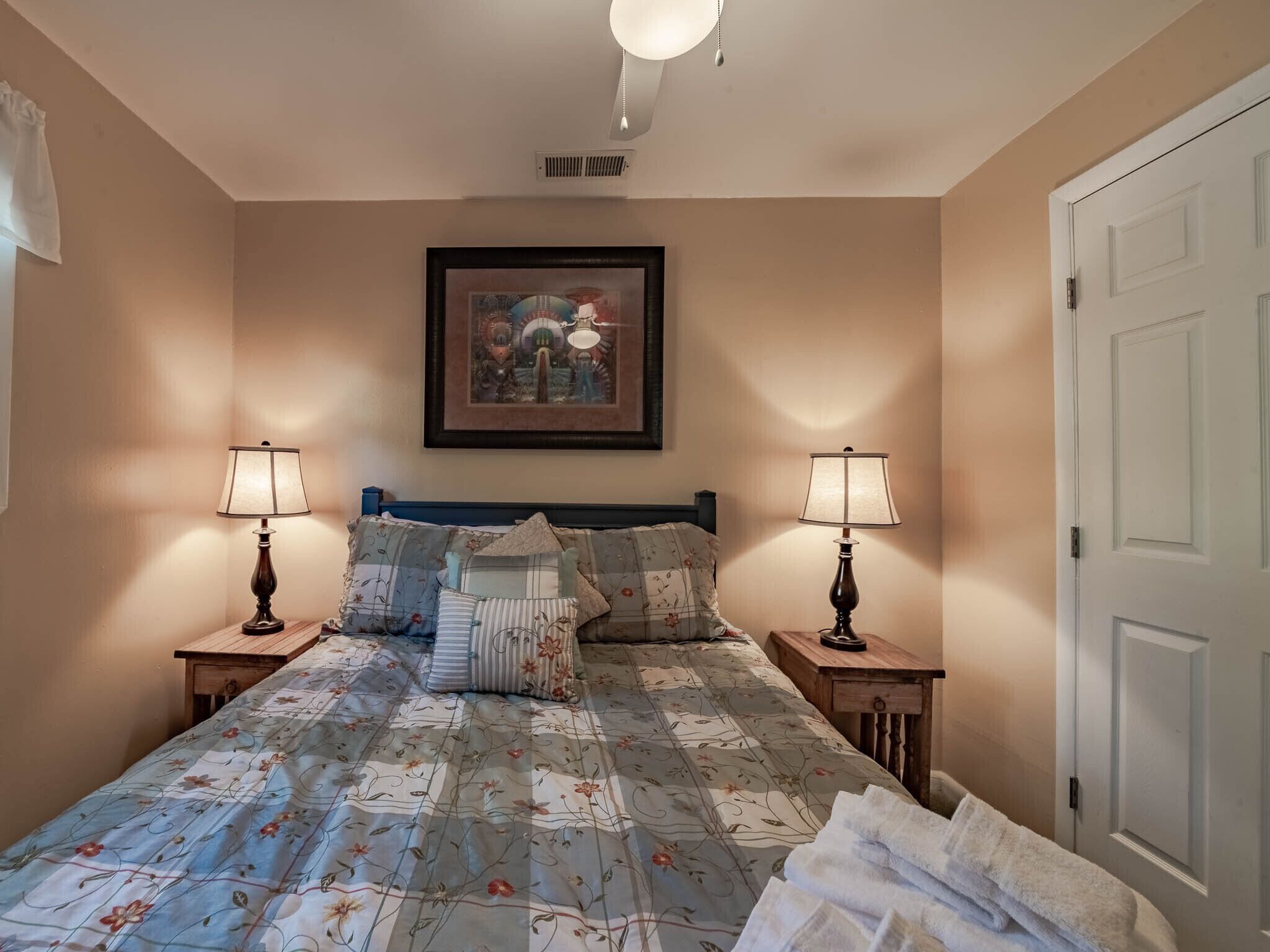 A bedroom with a bed, two bedside tables, a ceiling fan, a framed picture on the wall, and a door.