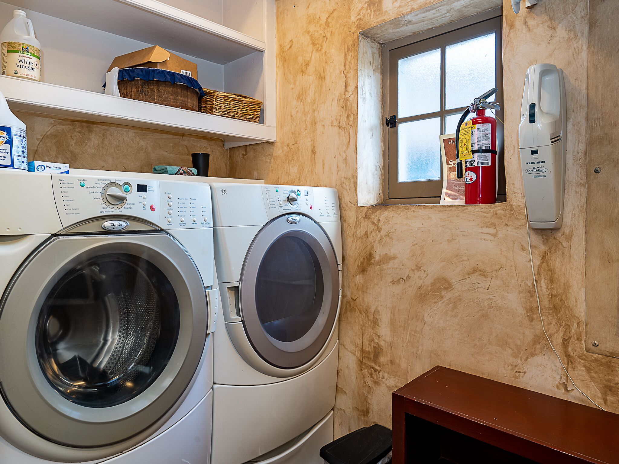 A laundry area with two front-loading washing machines, a shelf with cleaning supplies, a window with a view, and a fire extinguisher mounted on the wall.