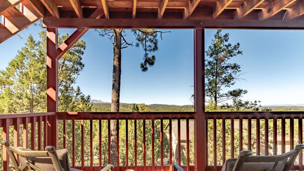 A covered outdoor seating area with two chairs and a table, overlooking a forested landscape. - Ruidoso Rabb by Ruidoso Vacation Rentals (Ruidoso)