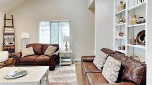 A spacious living room with two brown leather sofas, a white coffee table, and a white side table with a lamp. There is a bookshelf with decorative items and a window with blinds. - Ruidoso Rabb by Ruidoso Vacation Rentals (Ruidoso)