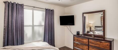 Habitación de hotel con cama, cómoda, televisor de pantalla plana, ventilador de techo y ventana con cortinas.
