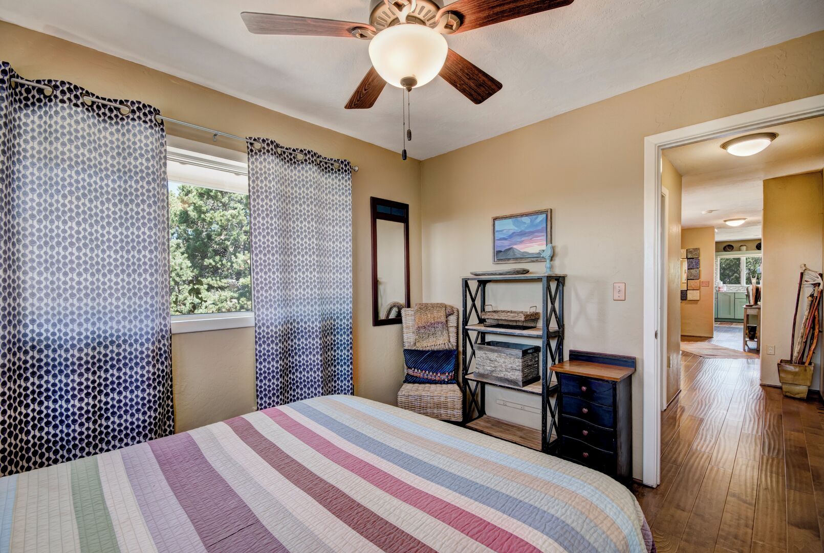 Quarto com cama, ventoinha no teto, janela com cortinas estampadas, uma cadeira, prateleira com cestos e vista para a cozinha.