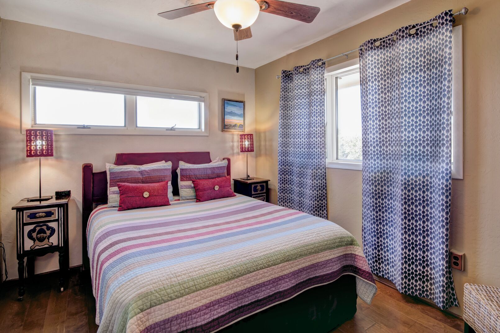 A bedroom with a bed, a ceiling fan, a window with curtains, and a bedside table with a lamp.