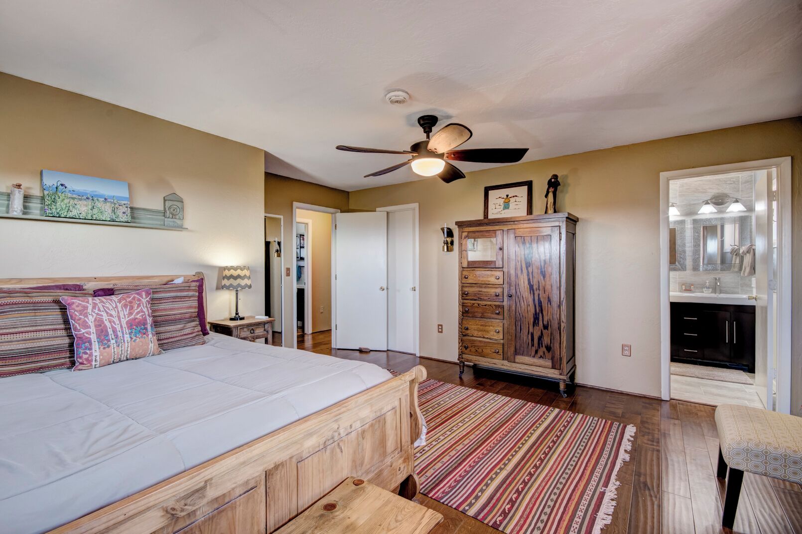 Une chambre à coucher avec un lit, une commode en bois, un ventilateur de plafond et une salle de bain visible à travers une porte ouverte.