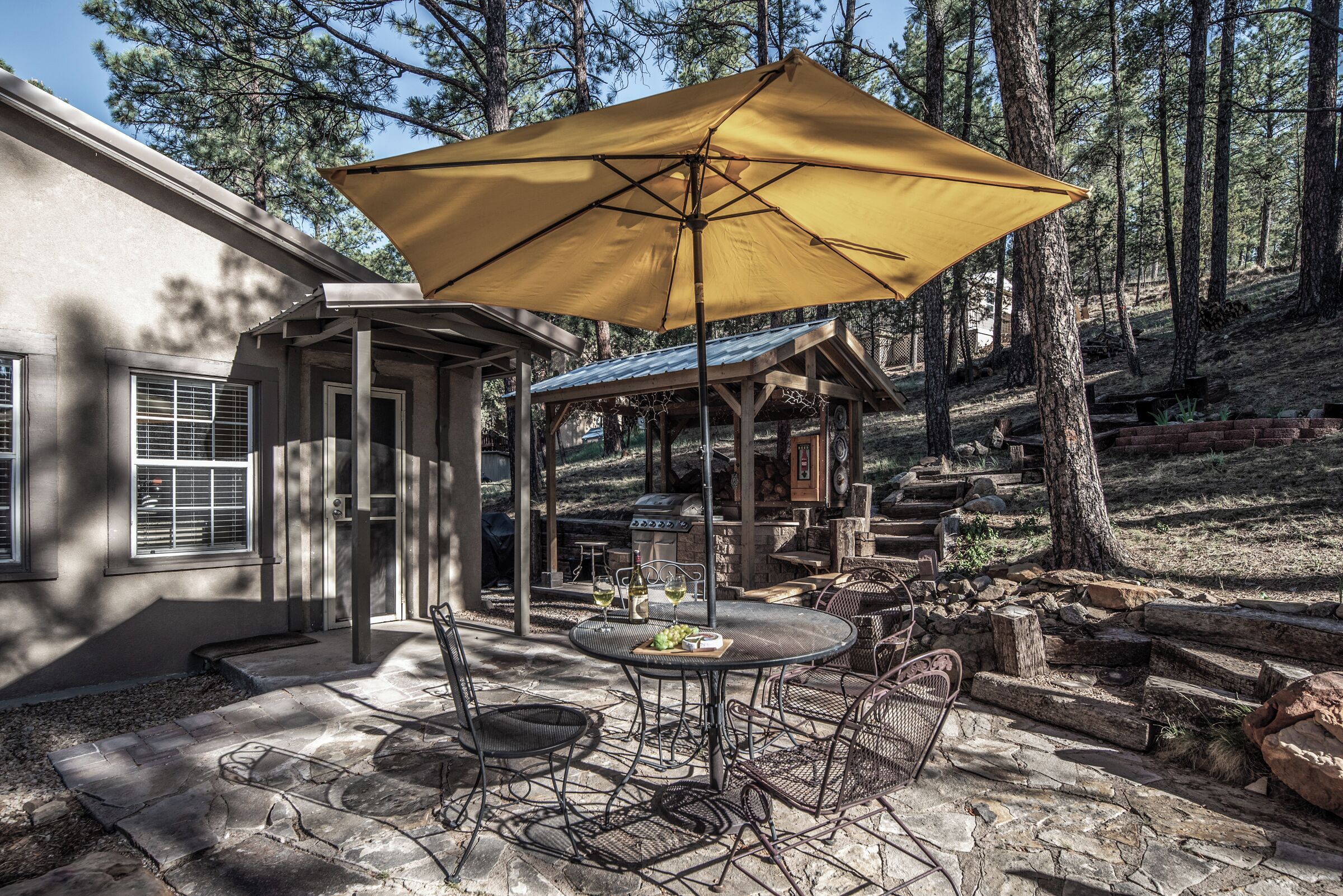 An outdoor patio with stone flooring, a round table, metal chairs, and a large yellow umbrella.