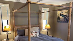Una cama con dosel cubierta con una colcha azul oscuro y almohadas a juego, flanqueada por dos mesitas de noche con lámparas, una alfombra estampada y un banco al pie de la cama.