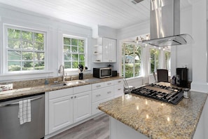 A modern kitchen with white cabinetry, granite countertops, a stainless steel range hood, and large windows offering views of greenery.