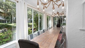 Une longue table en bois, entourée de chaises, se trouve dans une pièce lumineuse dotée de grandes fenêtres et d’un lustre en corde.