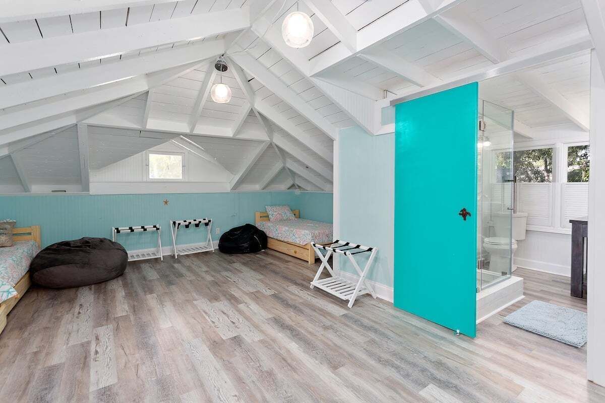 A spacious attic bedroom with a sloped ceiling, a teal door, and a large window.
