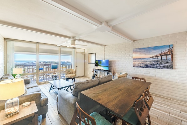 A spacious living area with a large wooden dining table, comfortable seating, a TV, and a scenic view through large windows.