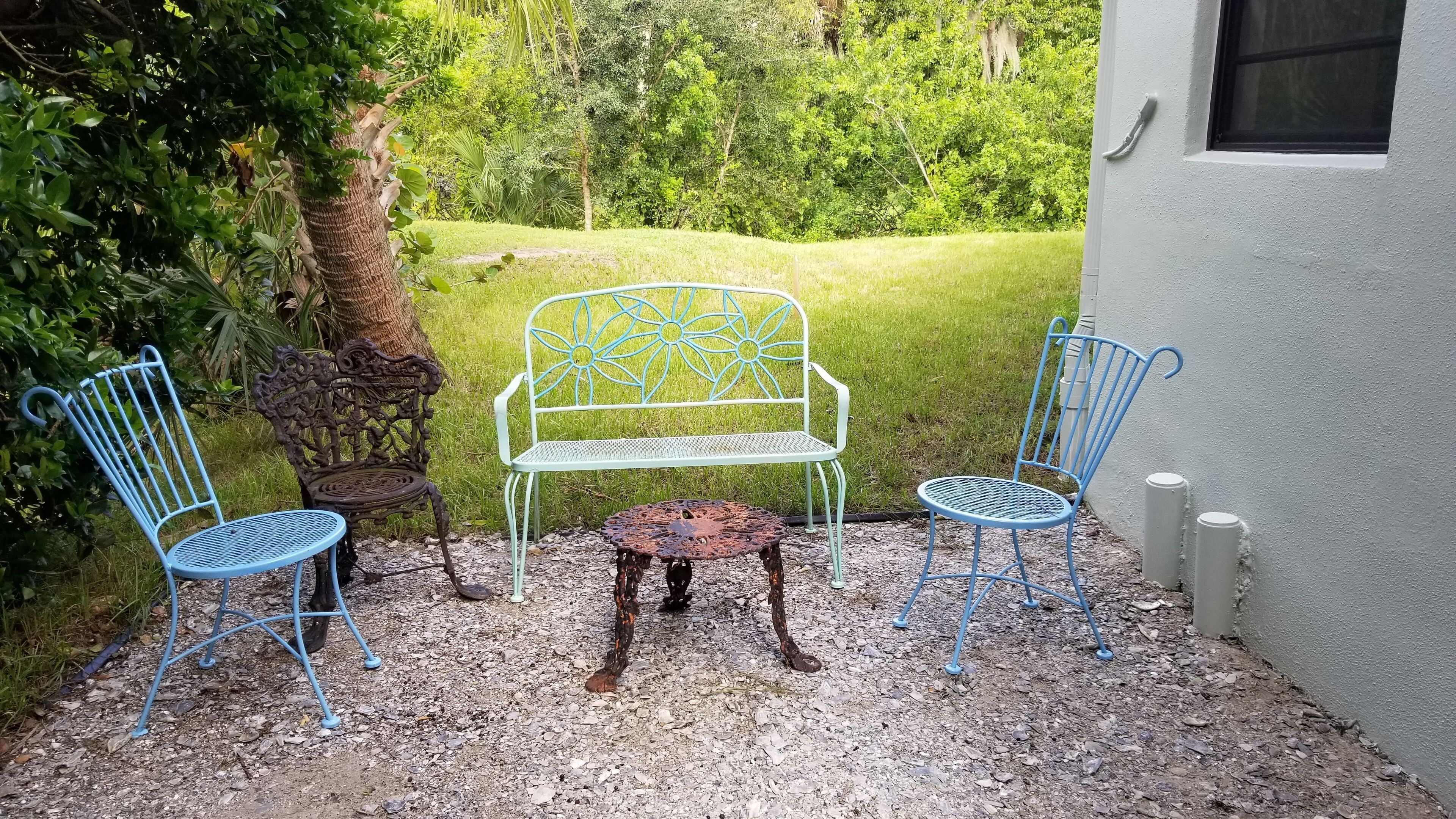 A patio with two blue metal chairs and a white metal bench with a floral design, surrounded by gravel and greenery.