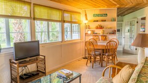 Un salon chaleureux avec un téléviseur posé sur un meuble en bois, une table basse en verre sur laquelle sont posés des livres, un canapé à motifs floraux et un coin repas avec des chaises et une table en bois.
