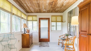Une pièce spacieuse avec un plafond en bois, de grandes fenêtres équipées de stores, une porte en bois, une table à manger avec des chaises et un buffet.