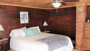 Een slaapkamer met een houten plafond, een bed met wit beddengoed en een grijze plaid, twee bedlampen, een nachtkastje en een bakstenen muur.
