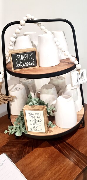 Un présentoir à trois niveaux avec des tasses en céramique blanche, une pancarte indiquant « BÉNÉDIT(E) SIMPLEMENT » et une autre pancarte posant la question : « COMMENT PRENDS-JE MON CAFÉ ? SÉRIEUX, TRÈS SÉRIEUX. »