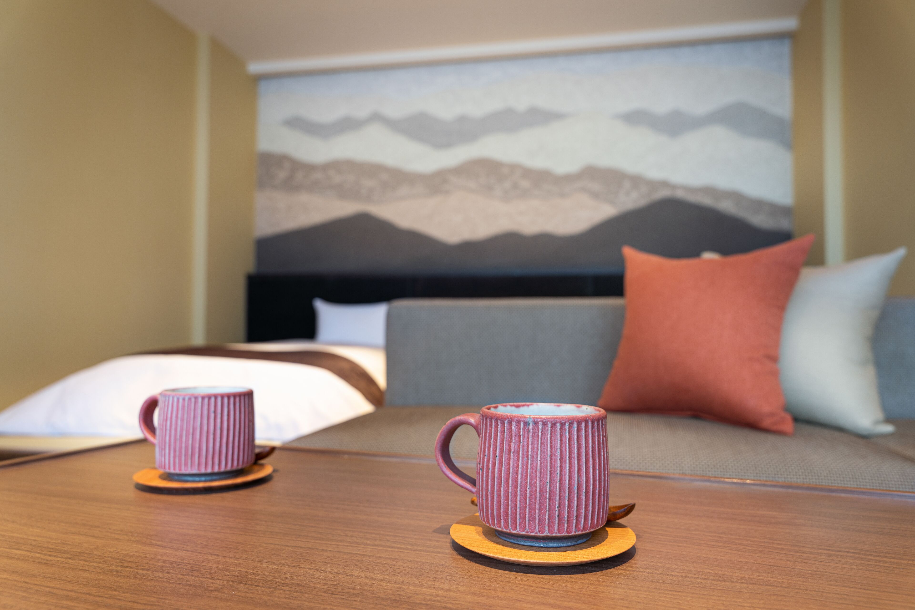 A hotel room with a bed, two mugs on coasters, a painting of mountains, and a sofa with pillows.