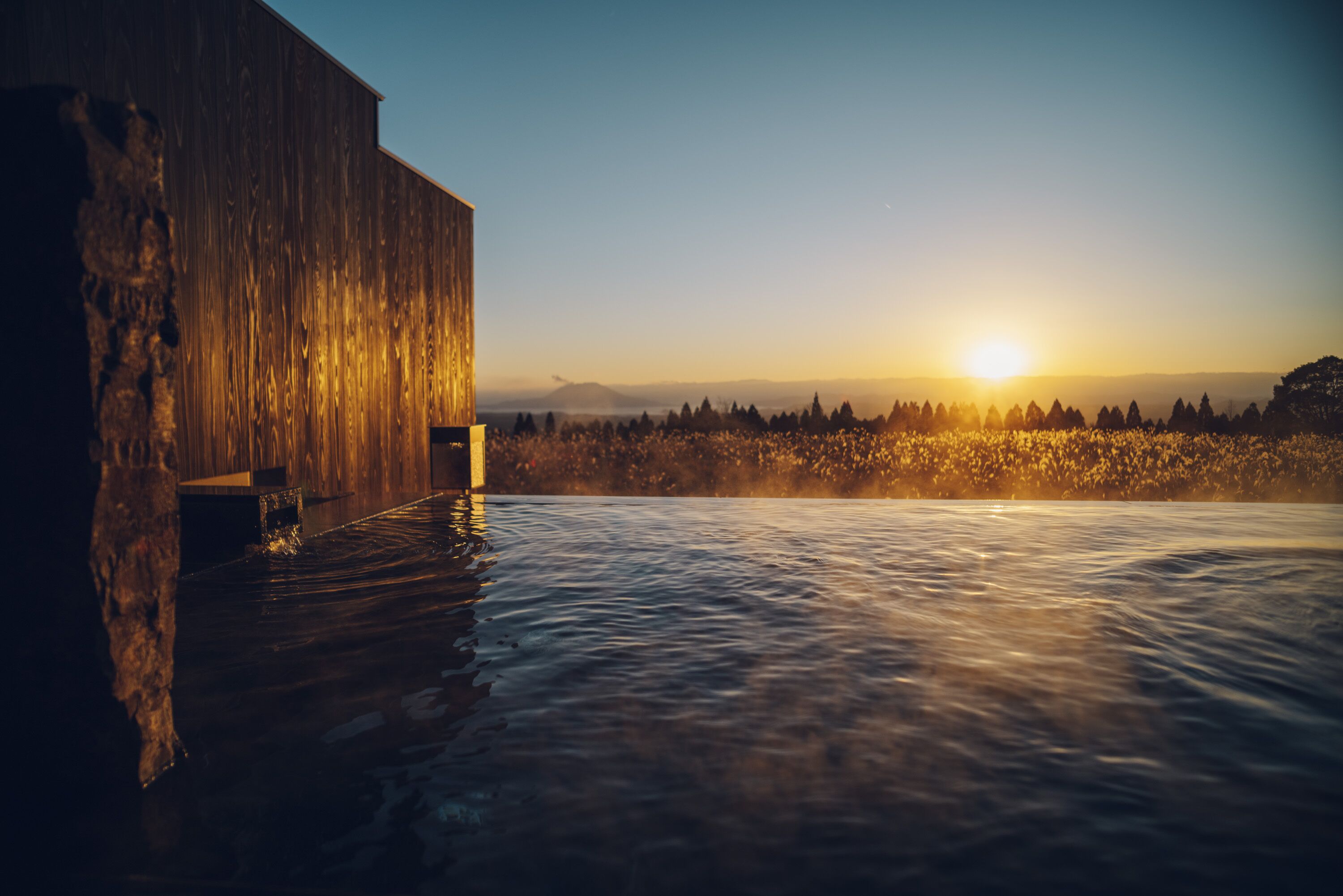A wooden building with a pool extending from its side, set against a backdrop of a sunset over a forested landscape.